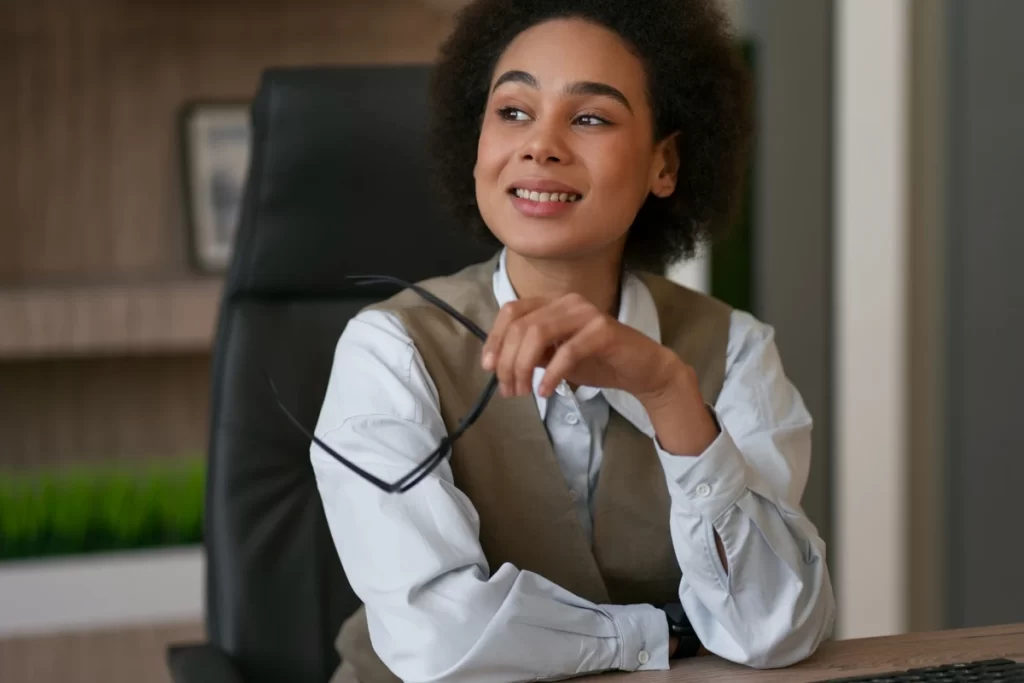 Mulher jovem com cabelo cacheado usando óculos e sorrindo em um ambiente de escritório moderno.