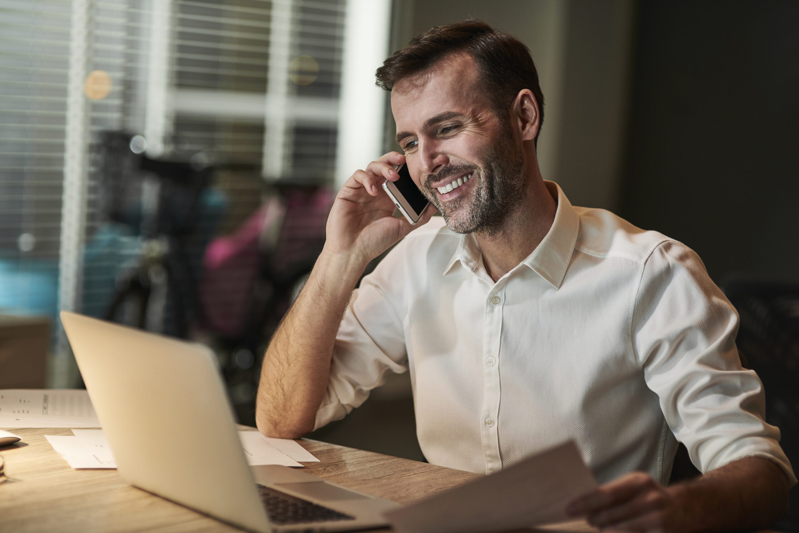 Empresario sorridente na frente de um computador falando no celular com roupa social.