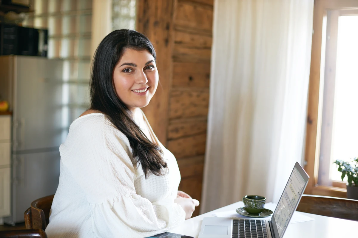 Mulher sorridente sentada em uma mesa de casa com laptop aberto, ambiente acolhedor com luz natural e decoração rústica.