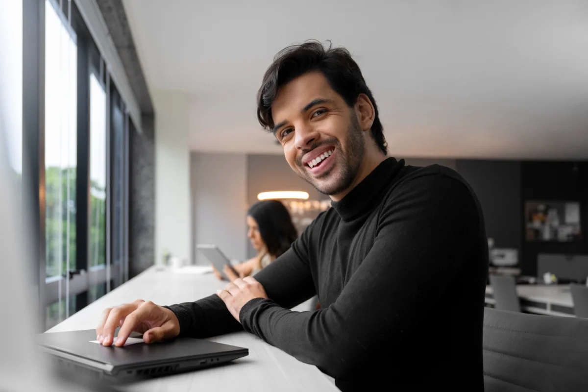 Homem sorridente usando suéter preto sentado em escritório moderno com laptop