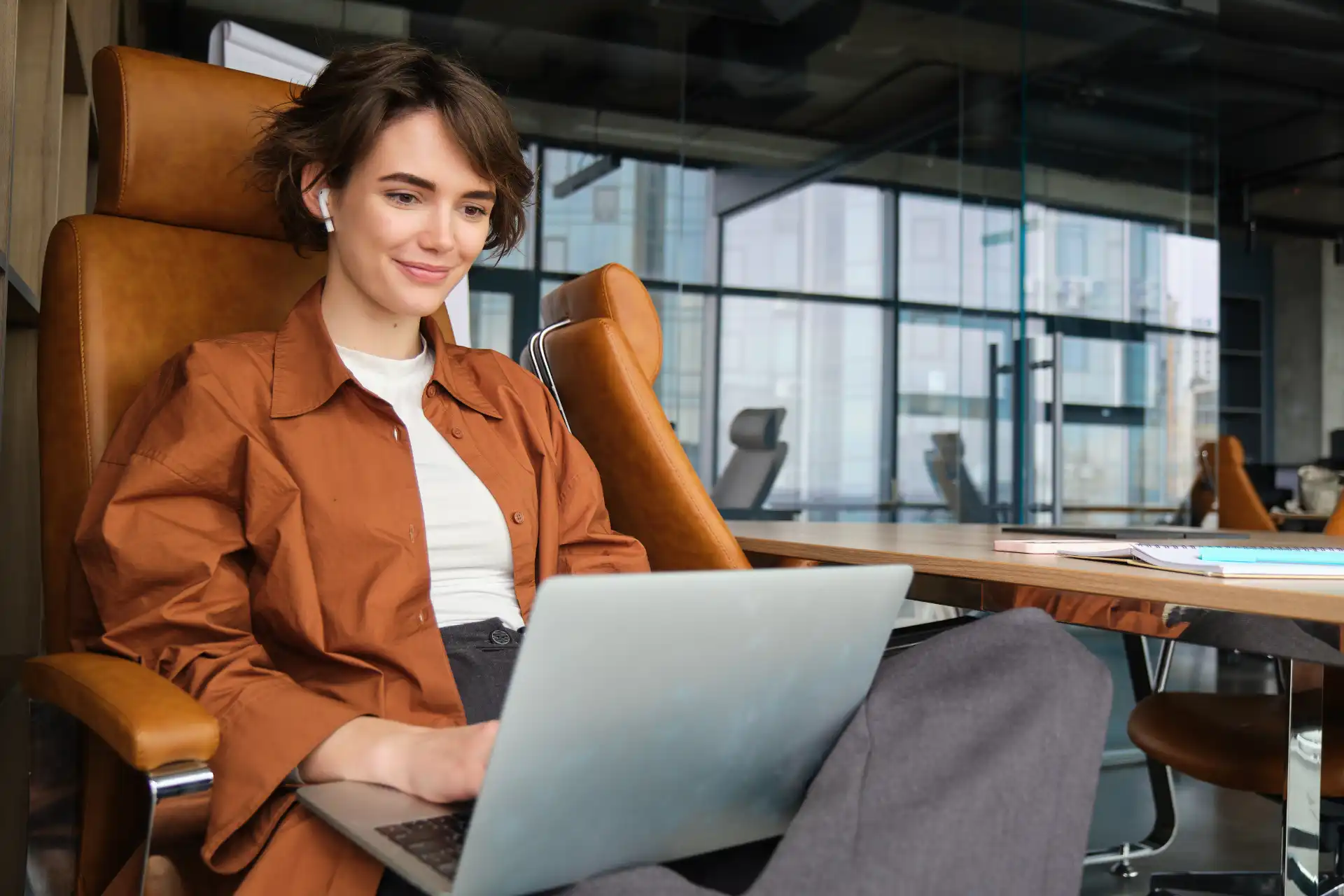 Mulher jovem de cabelo curto e sorridente, usando roupa casual marrom, trabalhando em um laptop em um escritório moderno com grande janelas de vidro.